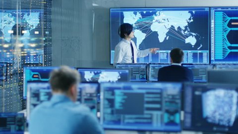 In the System Control Room Manager Holds a Briefing for His Staff Members. They're Work in Data Center and are Surrounded by Multiple Screens Showing Maps, Logistics Data. 