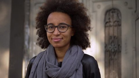 hipster young metis woman with dark curly hair wearing glasses standing in front of old building in city street, posing on camera with lovely smile slow motion
