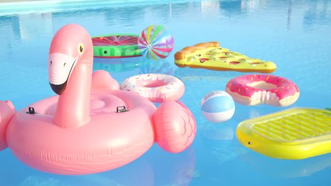 CLOSE UP: Empty inflatable flamingo, pineapple, pizza, doughnuts and watermelon floaties floating on pool water. Fun colorful floats waiting for pool party on hot summer vacation day.