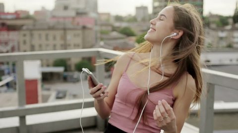 Music, technologies, VR, people, 4k and lifestyle concept - Girl listening music from smart phone mp3 player. at the sunny city streets and chatting with friends, sunset background