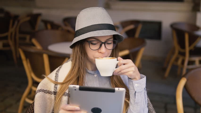Beautiful caucasian girl, in a grey striped hat, using tablet while having a cup of hot coffee, outdoor slowmotion