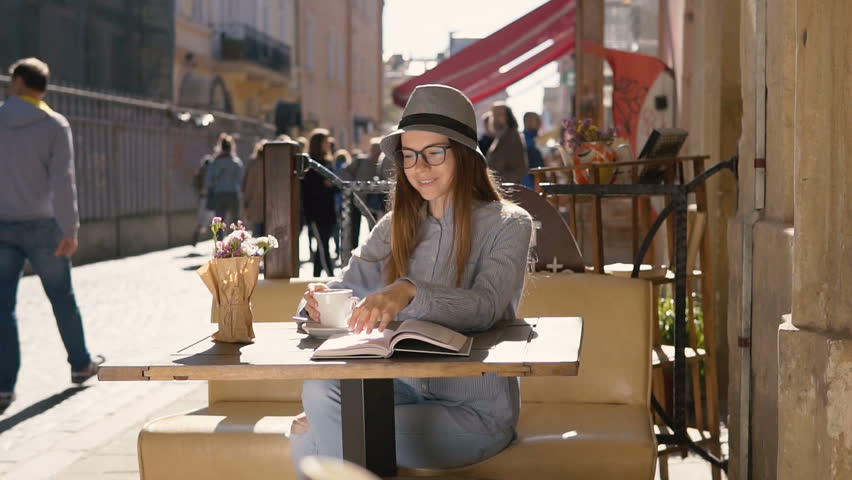 Smiling girl, in grey striped hat and casual outfit, enjoys coffee while reading in outdoor cafe, slowmotion on sunny day