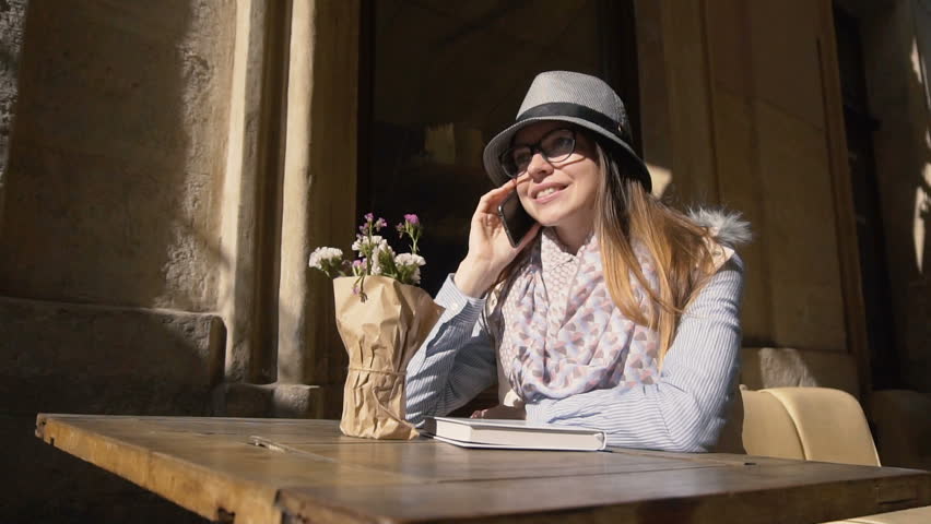 Caucasian girl, in grey striped hat and coloured scarf, chatting on the phone in outdoor cafe, slowmotion on sunny autumn day