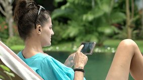 Woman watching video on smartphone and drinking water while relaxing next to the pool, steadycam shot
