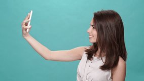 Happy brunette woman in t-shirt making selfie on smartphone over turquoise background
