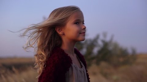 round footage of cute caucasian blondie girl wearing soft red cardigan standing in the middle of field, looking on camera enjoying wind blowing her hair early in morning slow motion 