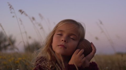 wind blowing hair of slavic cute little child enjoying sitting among flowers in meadow holding shell, listening noise from it with closed eyes feeling carefree on  sunset slowmotion closeup