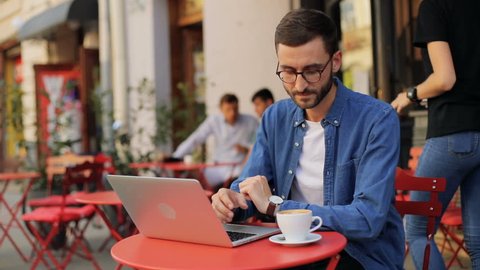 A closeup portrait of an attractive young man drinking coffe and then - working on his laptop at the cafe. Moving camera. Blurred background