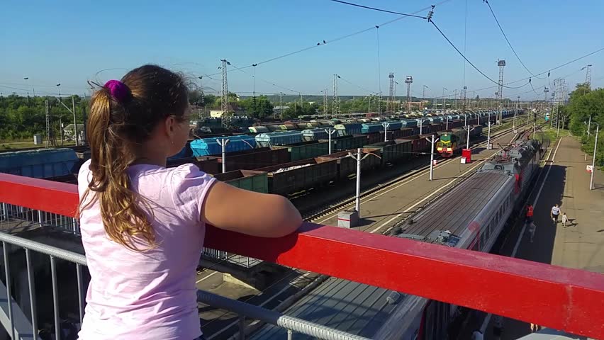 The girl is standing on the railway bridge at the station and looks afar at the trains, a shunting locomotive is passing under the bridge. Railways in Russia and Ukraine