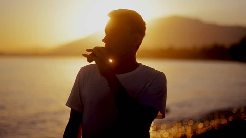 A guy at sunset smokes a e-cigarette. Vape. Vapping. Sea / Ocean / mounting / beach
The guy pours liquid into the electronic cigarette and smokes