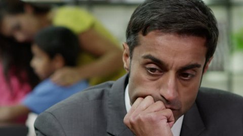 Close up of a man who is stressed about events in his life which are beyond his control. His wife is in the background, looking after their children