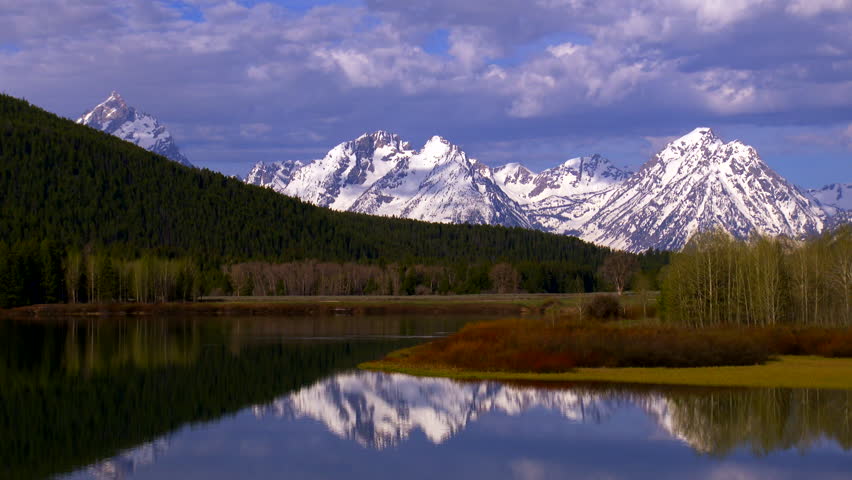 Grand Teton National Park - snow-capped mountain peaks at sunrise with dramatic lighting and cloud formations. Reflections in water.
