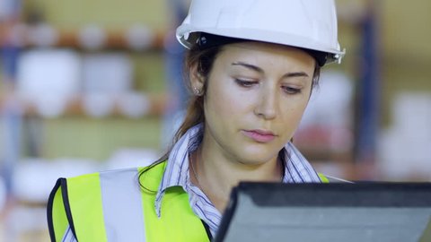 An attractive female warehouse employee wearing high visibility clothing and a hard hat is working on a digital tablet and checking her stock. In slow motion.