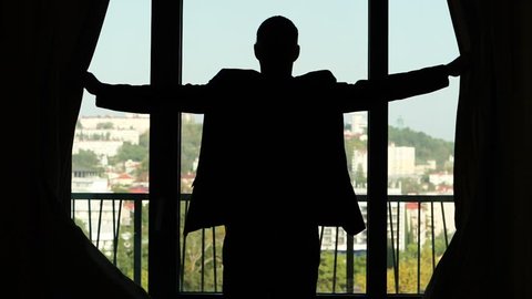Man expand both hands, open curtain, stand and look outside, sunny city seen blurred outdoors. Medium telephoto lens, half length view from back, black silhouette of guy in suit