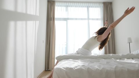 Woman sit on bed edge, stretch up hands and fall on soft blanket. Bright window, sun flash through transparent curtain, lazy weekend, relaxed morning time. Slide camera follow girl, slow motion shot