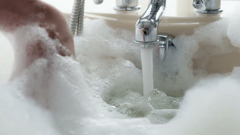 Bath is Filled with Fragrant Foam. The man's hand slowly sinks into a white foam against the background of a stream of water spurting from the tap. Filmed at a speed of 240fps