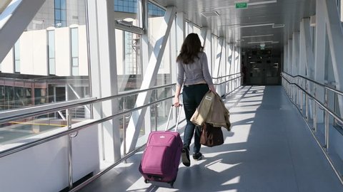 Passenger woman with trolley bag (large cabin luggage) walk through glass walled passage, come to airport terminal from airliner. Bright sun light and dark doorway ahead, follow camera move behind