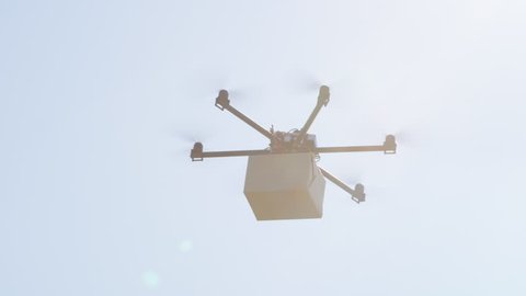 CLOSE UP UAV drone delivery. Multicopter flying big brown package over blue sky. Drone delivering post package to your home. Futuristic shipment by helicopter drone. Multirotor logistics and transport