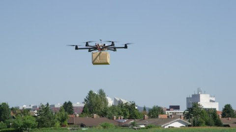 CLOSE UP: UAV drone delivery. Multicopter flying big brown package into city. Drone delivering post package to your home. Futuristic shipment by helicopter drone. Multirotor logistics and transport.