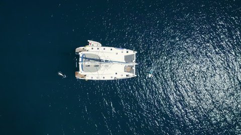Aerial Top Down Shot of a Big White  Sailing Yacht Anchored in a Bay with Man Standup Paddleboarding Near it. Ocean is Dark Blue and Beautiful. Shot on Phantom 4K UHD Camera. : vidéo de stock