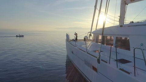 Aerial Side View Shot of Three Beautiful Fit Women Doing Morning Yoga on a White Sailing Catamaran Boat. People on the Boat Drive By. Peaceful Sea with Rising Sun and Calm Sea. Shot on Phantom 4K UHD