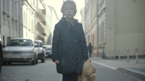 Sad little girl with teddy bear staying in the street, turning and go away.