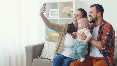 family, parenthood and people concept - happy mother and father with baby taking selfie by smartphone at home