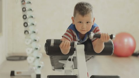 Smiling Young boy doing heavy dumbbell exercise for biceps. Baby working out with dumbbells on training apparatus. Strength and motivation. The concept of a healthy lifestyle. GYM