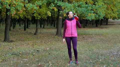Beautiful fitness sport girl in autumn park in sportswear drinks water or isotonic drink from a sports bottle.
