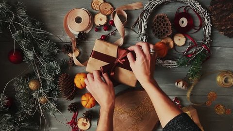 A top-down plan. Fully visible the table with the decorations. Female hands put and finalize Christmas gift wrapped in craftool paper on a wooden table. Bandaging tape and tied in a bow.