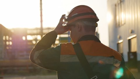 slow motion backside view skilled worker in outfit walks along huge refinery territory under sunshine