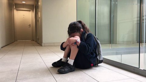 One sad young elementary school girl (female age 5-6) wearing school uniform and backpack sitting on a corridor floor of a building and crying. Childhood and education concept. Real people. Copy space
