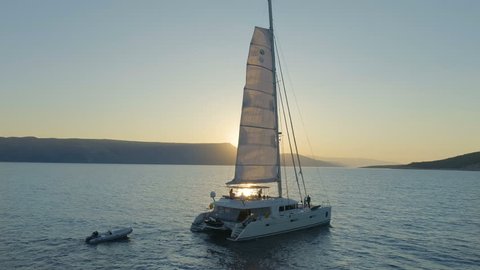 Aerial Shot of a Sailing Catamaran Yacht with Raised Sail Traveling Through The Calm Seas with Sun Rising Behind the Hills. People Relaxing on the Deck.