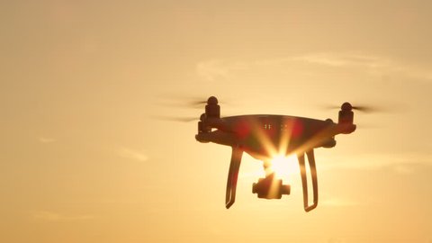 CLOSE UP LENS FLARE SILHOUETTE Small filming drone flying over golden sunset sky. White quadcopter drone with attached camera filming at golden light. Quadrocopter helicopter flying across sunrise sky