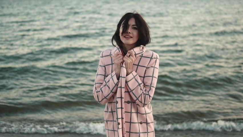 Portrait of an attractive young woman in a coat on the beach. girl smiling, posing and looking at camera. Slow motion