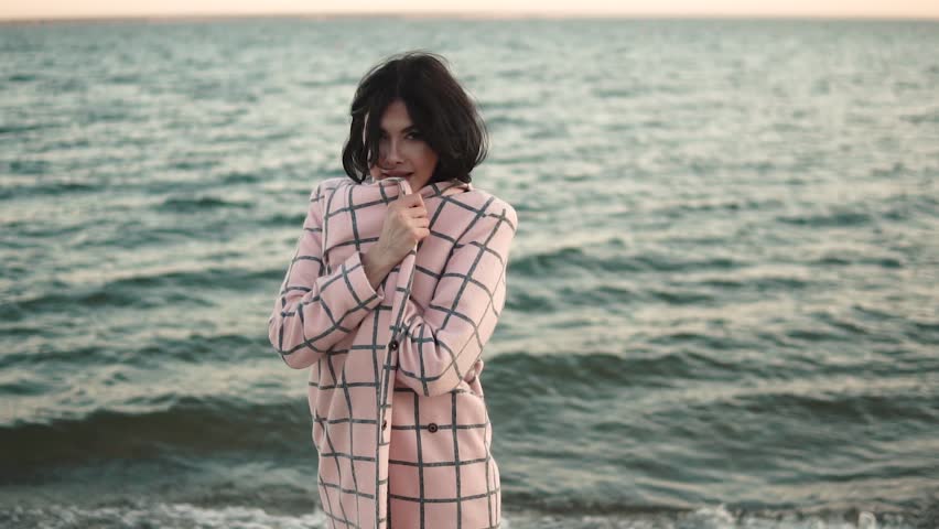 Young woman in elegant coat walks on the beach. The girl is enjoying the warm autumn day. Portrait of beautiful girl near the water. Slow motion