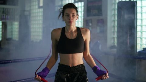 Young female boxer with her hands wrapped in bandage jumping on the skipping rope in a gym with smoke. Slowmotion shot