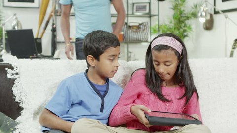A young boy and girl of Indian ethnicity are playing on a digital tablet computer together at home and their father comes to see what they are doingの動画素材