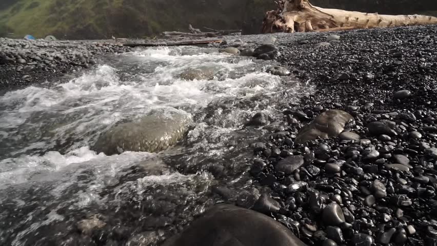 Dolly shot of a freshwater stream flowing into the ocean on a rocky beach. Camera moving upstream towards driftwood tree