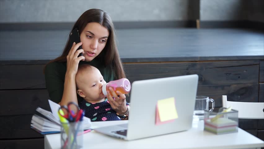 Young mother is giving juice to her baby from bottle and speaking by telephone. A woman on maternity leave is working from home, doing important phone call, holding baby, sitting in front of laptop. | Shutterstock HD Video #31889044