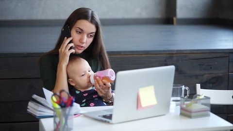 Young mother is giving juice to her baby from bottle and speaking by telephone. A woman on maternity leave is working from home, doing important phone call, holding baby, sitting in front of laptop.