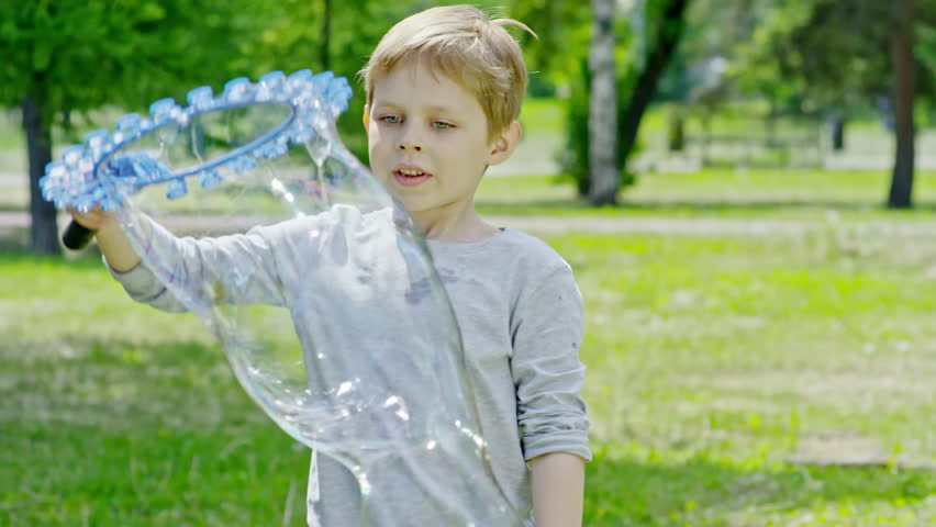 Little boy swinging wand and making huge long soap bubble and lots of small bubbles while playing in the park