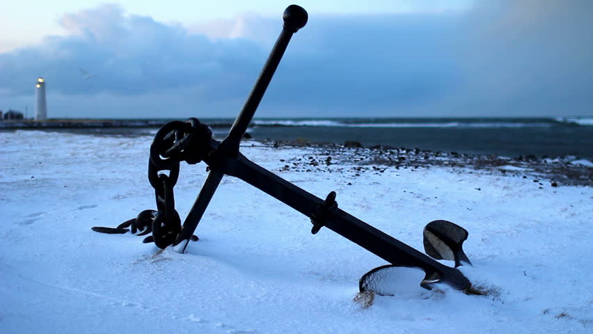  A ship anchor laying next to the ocean with a lighthouse in the background. Reykjavik, Iceland