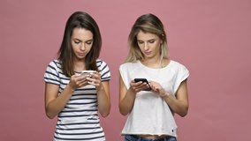Two serious concentrated caucasian women using smartphones and typing isolated pink