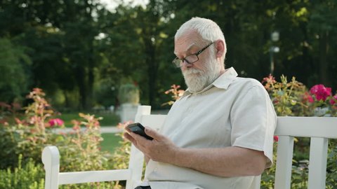 An old man sitting on a white bench in the park and using a phone. Medium shot. Soft focus