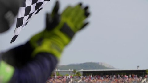 Man holding and waving Checkered race flag in slow motion at finish line on a raceway. Hands of unidentified person applauding.