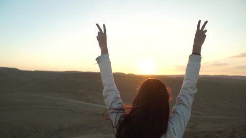 Tilt down on a young woman with her arms up in victory in a desert scenery. Success and happiness over a win or positive outcome. Freedom and carefree feeling in nature. Happy cheerful doing v sign