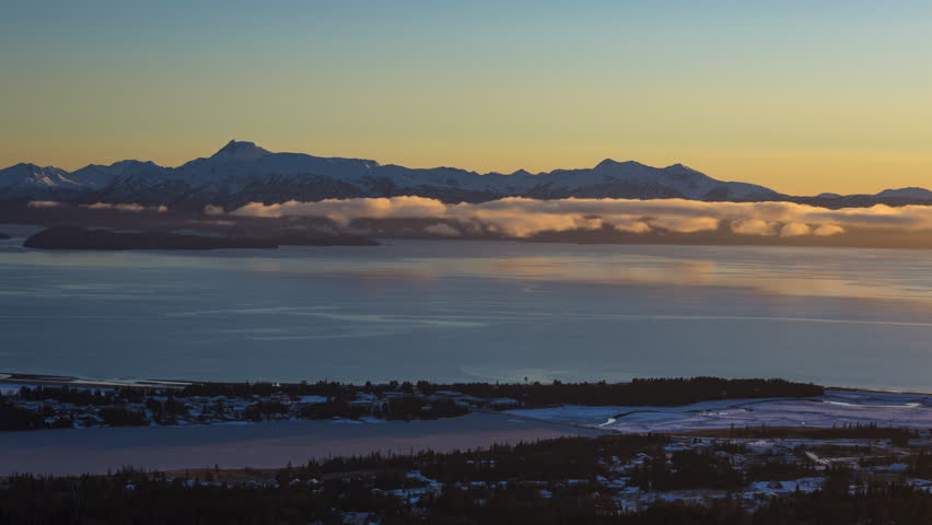 A time-lapse of the setting sun over Homer, Alaska with Beluga Lake in the foreground, rolling clouds in the center, and snow capped mountains in the distance