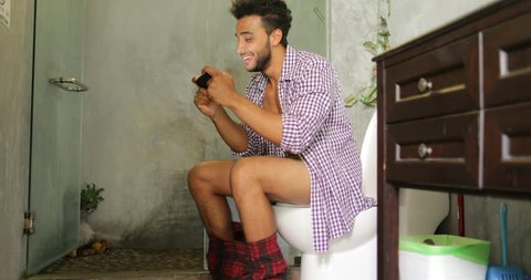 Man Sitting On Toilet Use Cell Smart Phone Young Latin Guy Playing Game In Restroom
