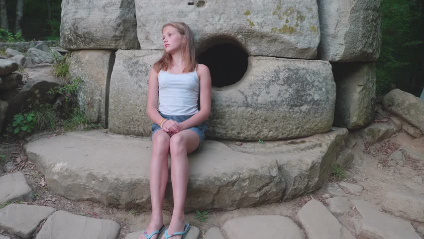 Young caucasian girl pensively sits at an ancient dolmen